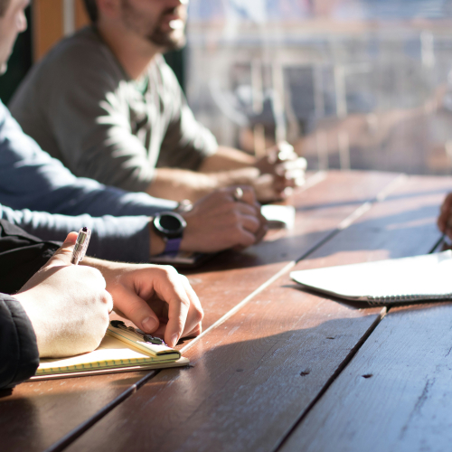 A group of people sitting around a wooden table in a meeting, writing notes in notebooks and discussing ideas in a bright, informal setting.