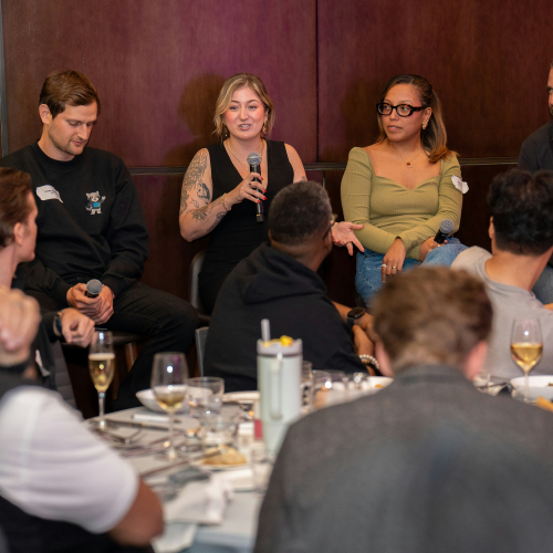 Four speakers take part in a panel discussion with microphones while an audience seated at tables listens during a conference-style event