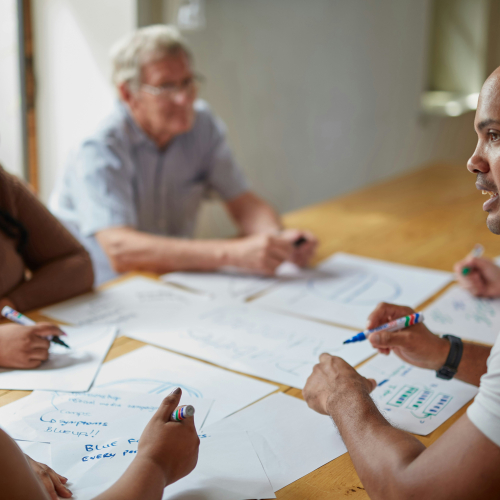 A diverse group of people gathered around a table, actively discussing and writing ideas on large sheets of paper, showing collaborative planning and teamwork.